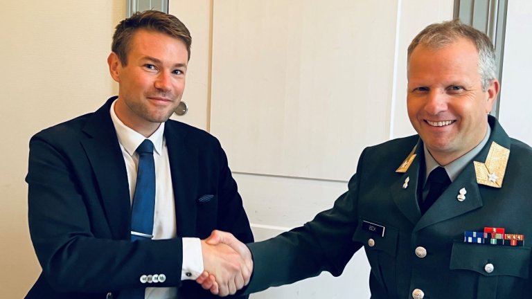 To smilende menn tar hverandre i hånden, den ene ikledd dress og slips og den andre i militær uniform.