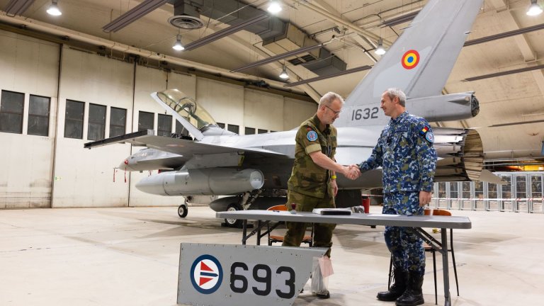 To menn i militær uniform tar hverandre i hendene inne i en flyhangar, med et F-16 jagerfly i bakgrunnen.