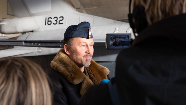 Jagerflyver i frakk med pelskrage blir intervjuet av pressen foran et F-16 jagerfly.