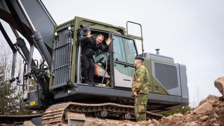 Svartkledd mann vinker fra døråpningen på en gravemaskin, en uniformskledd mann ser smilende mot ham.