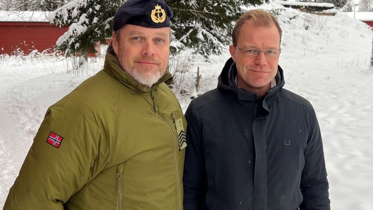 En militært kledd og en sivilt kledd mann ser inn i kameraet foran en snødekket gran