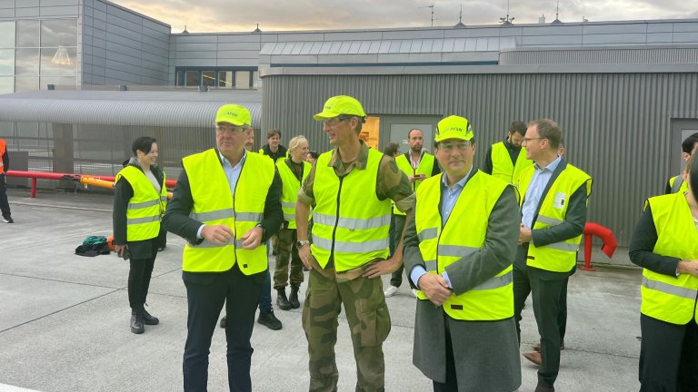 Tre menn i gulevester og gule hjelmer står og snakker sammen foran en flyplassbygning.