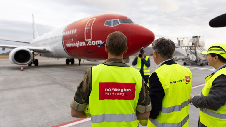 Tre menn med gule vester står med ryggen mot fotograf og ser mot et Nowegian-fly på oppstillingsplass