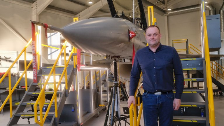 Blåkledd mann står foran et jagerfly inne i en hangar.