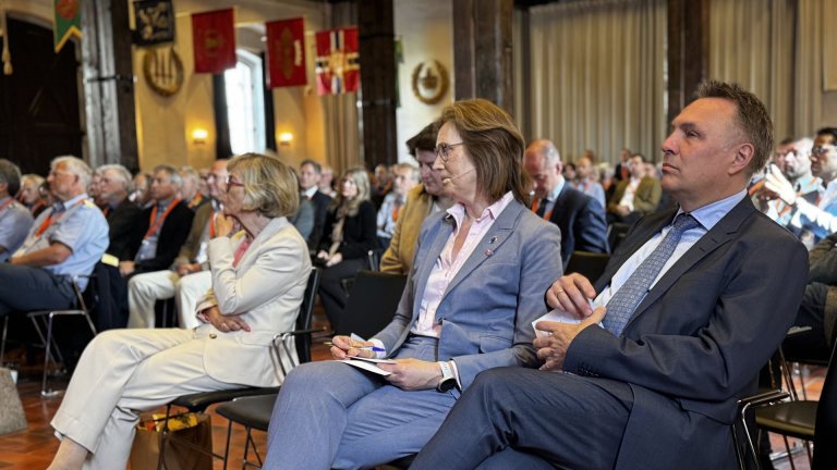Deltakere sitter i salen og følger med under en konferanse, med fokus på tre personer i forgrunnen som lytter og noterer.