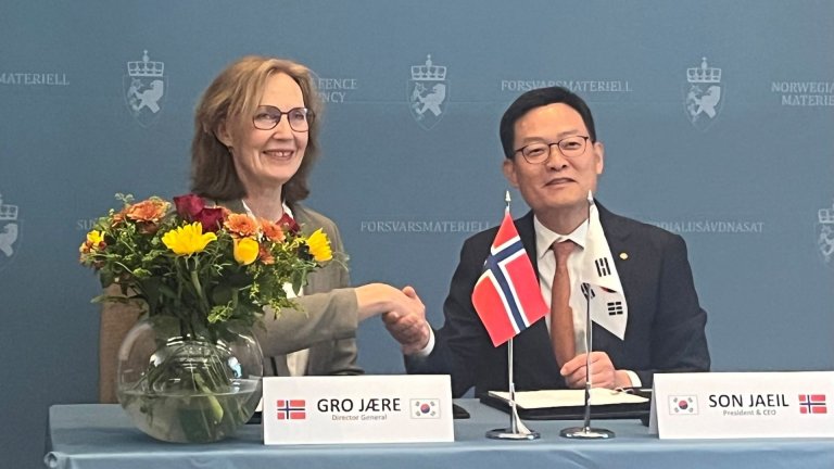 Kvinne og mann smiler og håndhilser under en signeringsseremoni med norske og sørkoreanske flagg på bordet.