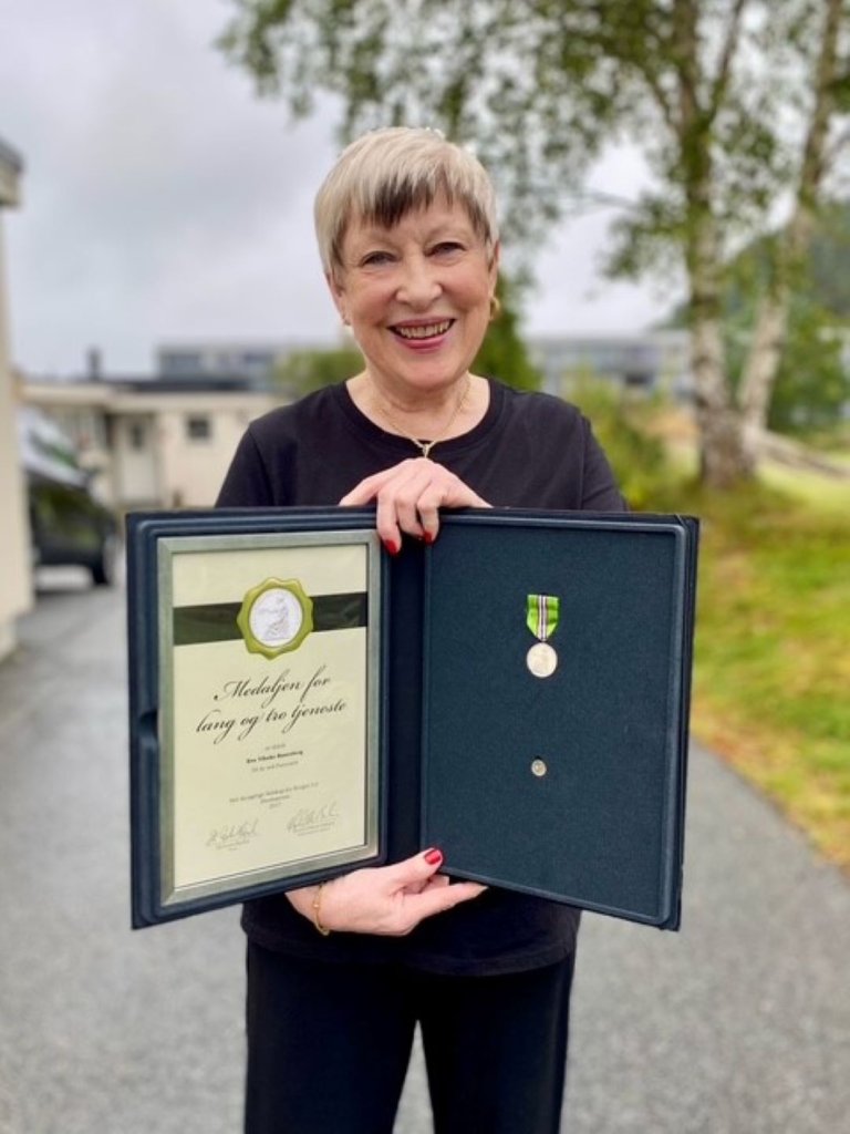 En smilende kvinne står utendørs og holder en åpen eske med medalje og diplom. Diplomet viser at hun har mottatt Medaljen for lang og tro tjeneste. Hun er kledd i svart og står på en gruslagt vei med gress og trær i bakgrunnen