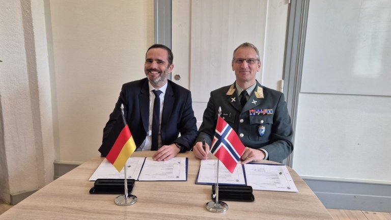 To menn sitter ved et bord foran hver sin nasjonsflagg, Tyskland og Norge, og har nettopp signert dokumenter. Den ene er i dress og den andre i militæruniform med medaljer og distinksjoner. Begge smiler mot kameraet