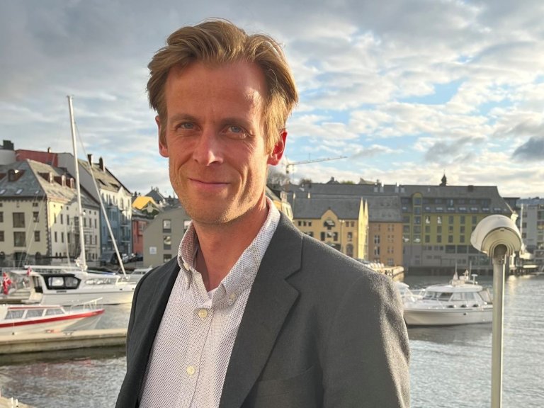 Mann i dressjakke smiler mot kamera foran en havn med båter og bygninger i bakgrunnen.