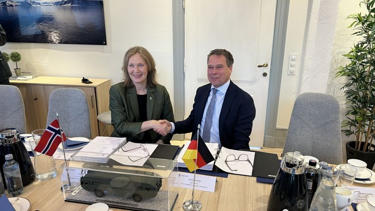 Kvinne og mann ved konferansebord gir hverandre et håndtrykk. Det står et norsk og et tysk flagg på bordet.