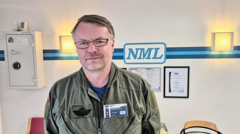 Mann i uniform med navneskilt foran en vegg merket med logoen "NML"