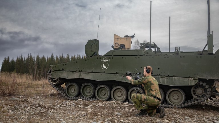 Soldat med hodetelefoner og holder en drone ved siden av et militært beltekjøretøy.