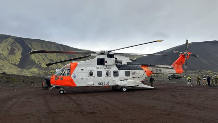 Redningshelikopter parkert i vulkansk landskap med fjell, mens personell beveger seg rundt.