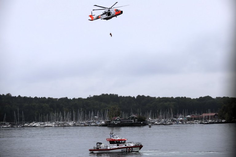 Redningshelikopter heiser en person mens en redningsskøyte ligger i vannet under, med en båthavn i bakgrunnen.