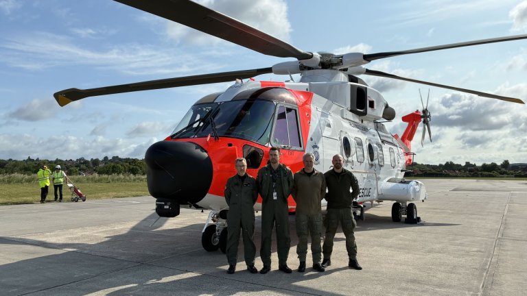 Fire personer i uniform står foran et norsk redningshelikopter på en flyplass, med blå himmel og lette skyer i bakgrunnen.