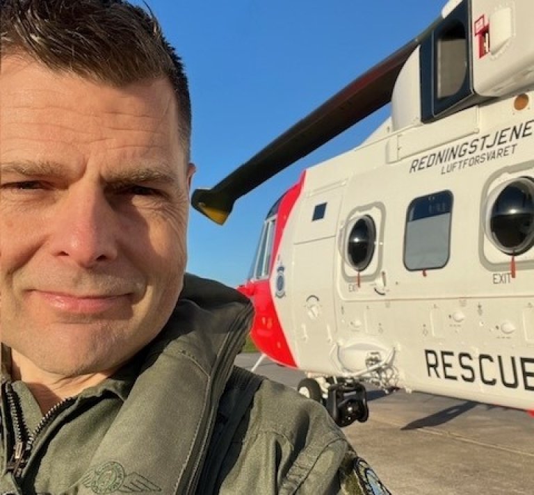 En mann i flyuniform tar en selfie foran et redningshelikopter.