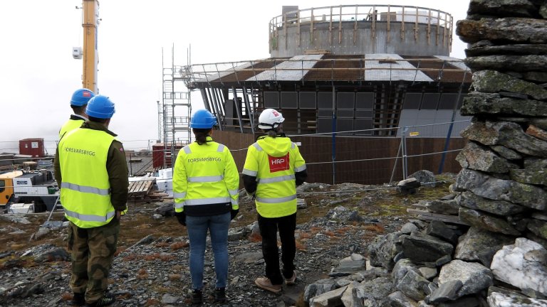 Fire personer med verneutstyr og refleksvester ser mot et radarbygg under oppføring i fjellet.