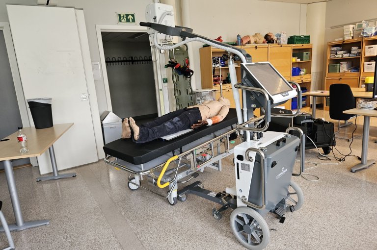 Et undervisningsrom for medisinsk trening, der en realistisk øvingsdukke ligger på en båre. Ved siden av står avansert medisinsk utstyr, inkludert en skjerm og en mobil enhet på hjul. Rommet er fylt med hyller, stoler og annet undervisningsmateriell