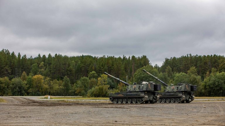 To kamuflasjemalte artillerikjøretøy står på en grusplass med skog i bakgrunnen.