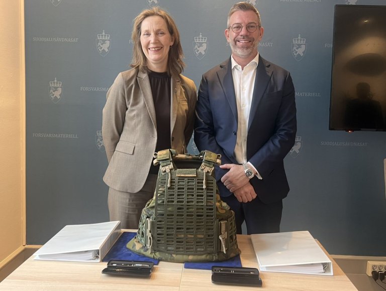 To personer står ved et signeringsbord med en kampvest stående på