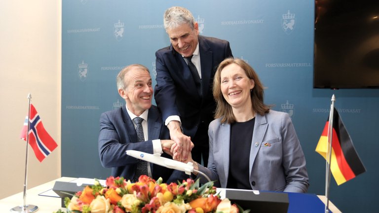 Tre smilende mennesker tar hverandre i hånden, sittende ved et bord med norsk og tysk flagg og en liten modell av et missil.