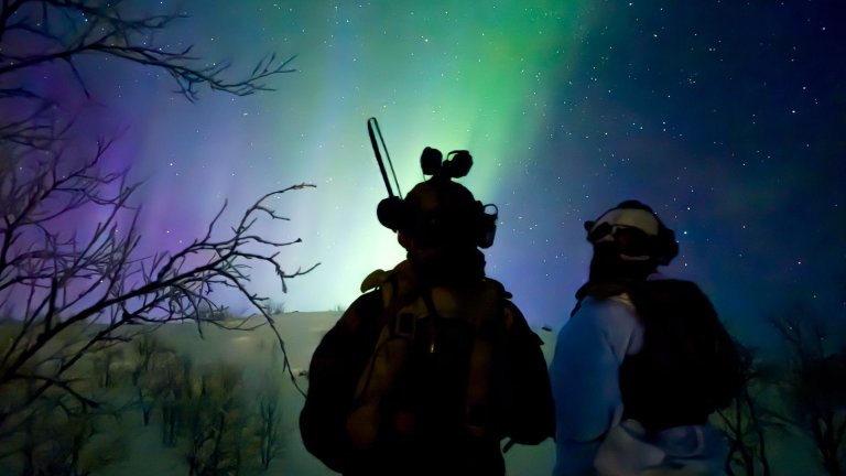 To vinterkledde soldater ser opp mot himmel med nordlys