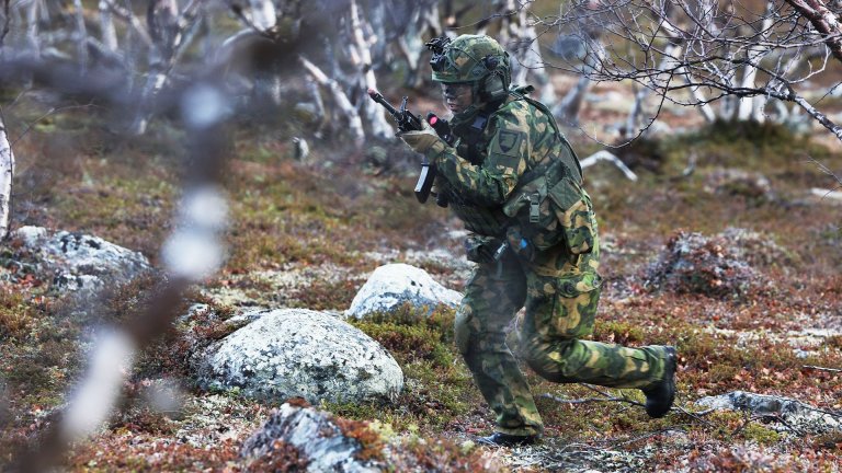 Soldat i kamuflasjeuniform løper med våpen gjennom skogterreng