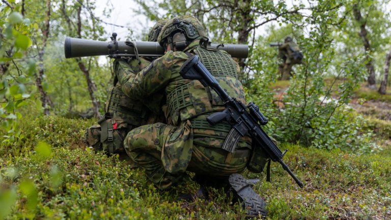 To soldater i kamuflasjeuniform sitter i skogen, én av dem sikter med et skulderavfyrt våpen
