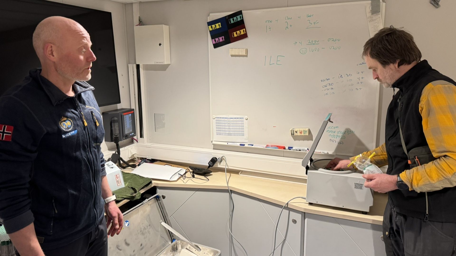 To menn i et rom med laboratorieutstyr. Den ene mannen holder en pose med prøver.
