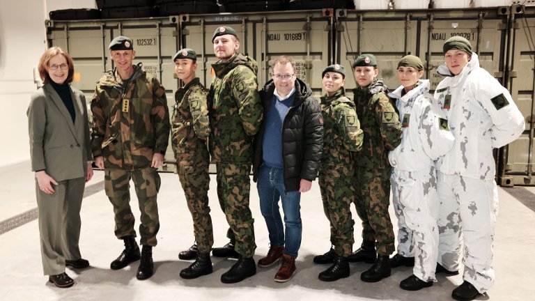 En gruppe soldater og to sivilt kledde personer poserer foran militærcontainere innendørs